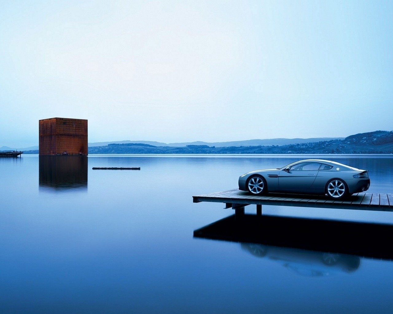 Silver Aston Martin Zagato V8 Vantage parked on a wooden pier overlooking a calm blue lake beneath a pale sky.