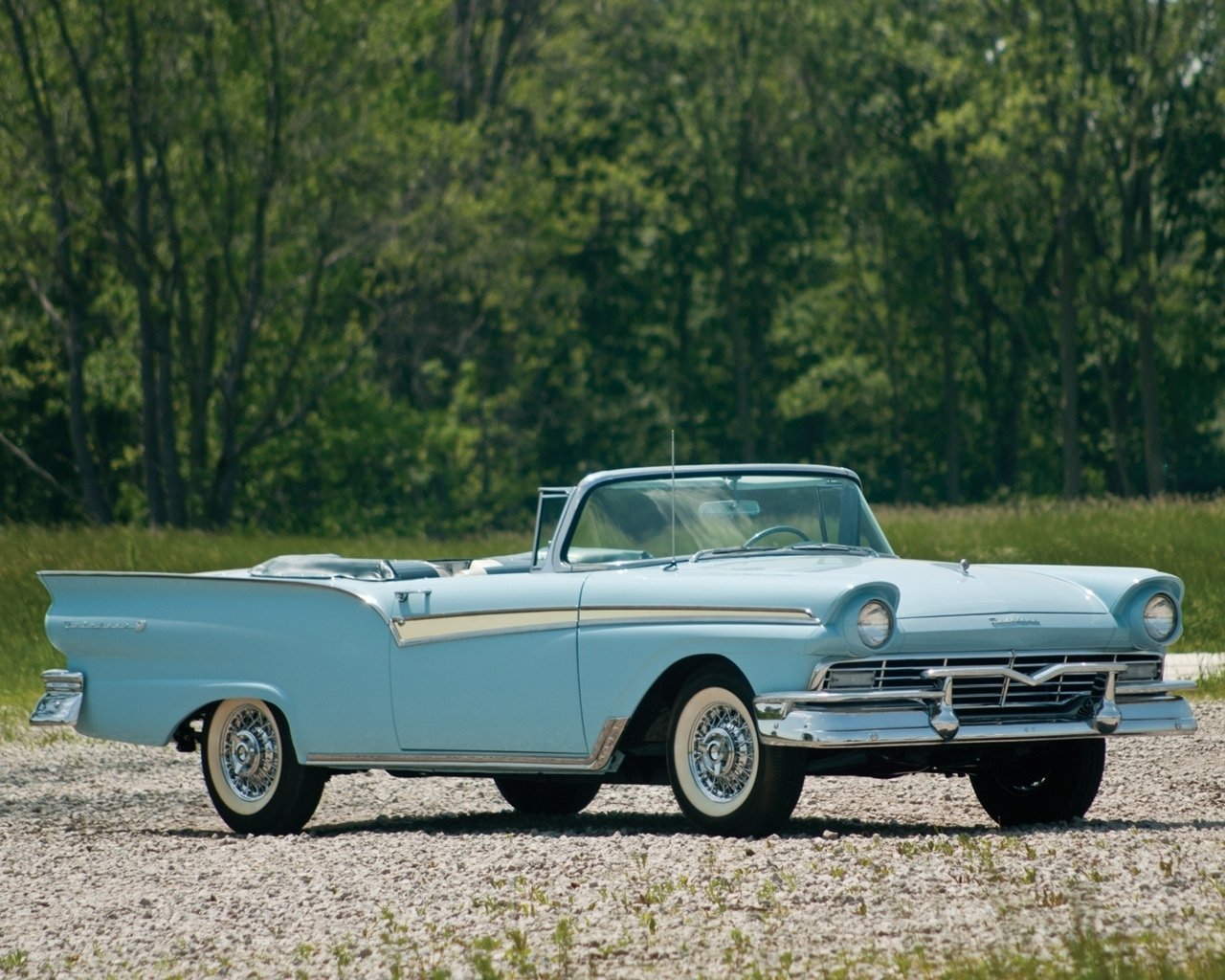 Classic light-blue convertible vehicle parked on gravel with a green treeline in the background.