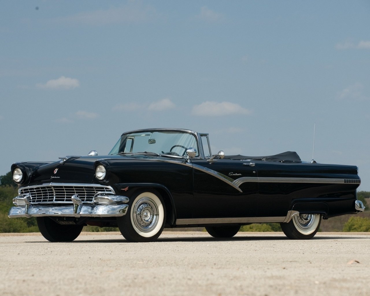 Black classic convertible vehicle with chrome trim and whitewall tires parked on open pavement under a clear blue sky.