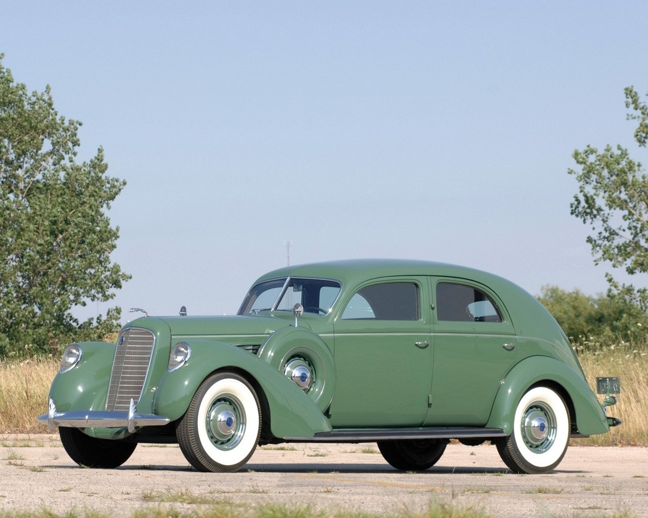 Classic green vehicle — a 1930s streamlined sedan with whitewall tires and chrome grille, parked on a gravel road beside open fields.