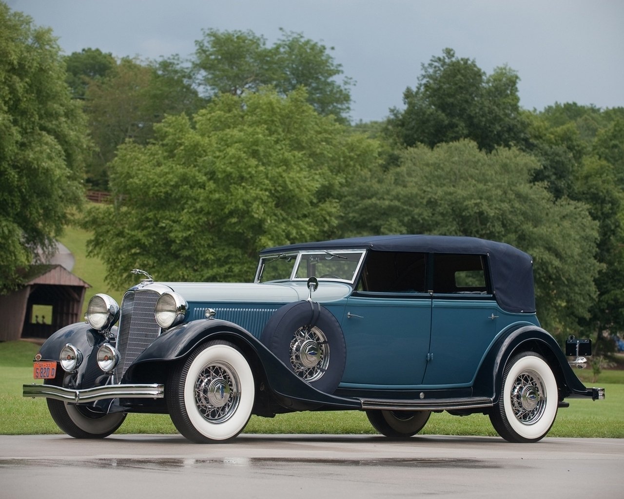 Classic vehicle: blue vintage convertible with soft top, chrome accents and whitewall tires parked in front of a tree-lined park.