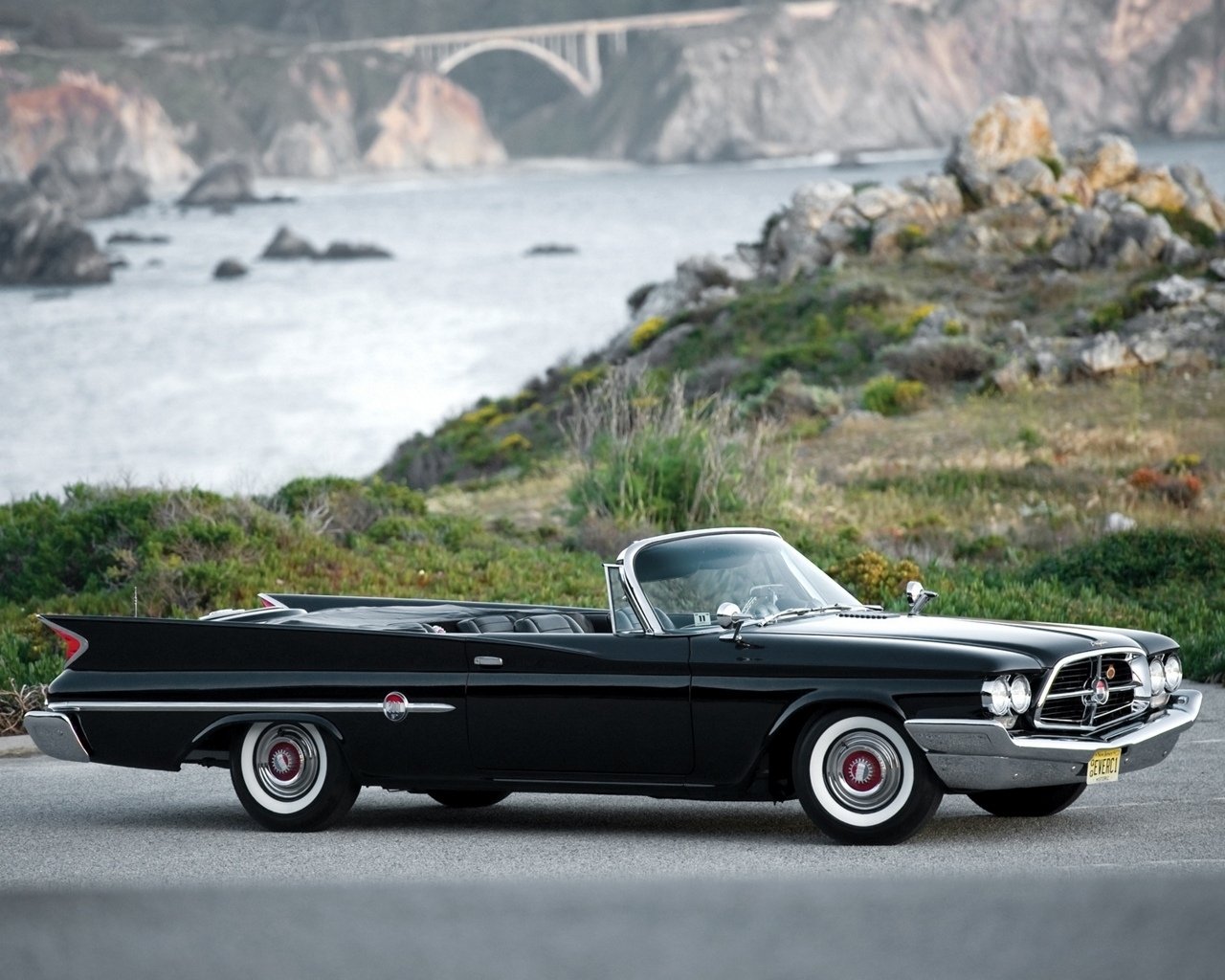 Black classic convertible vehicle parked on a coastal road, rocky cliffs and an arched bridge visible in the misty background.