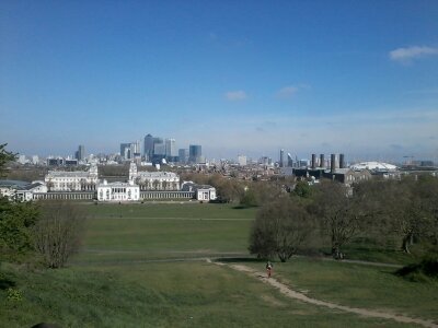 Green park and dirt path lead to historic waterfront buildings with the man-made London city skyline on the horizon beneath a clear blue sky.