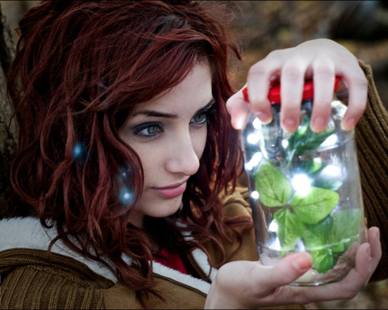 Susan Coffey gazes intently at a jar filled with glowing lights and green leaves, set against a blurred natural background.