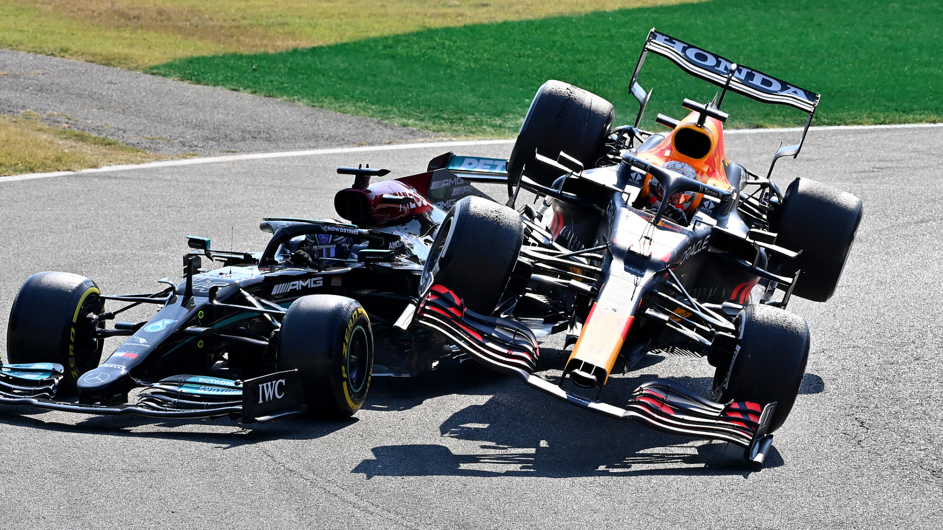 Two F1 cars side-by-side in an intense motorsport battle, one briefly airborne over a kerb as they jostle for position on a sunlit racetrack.