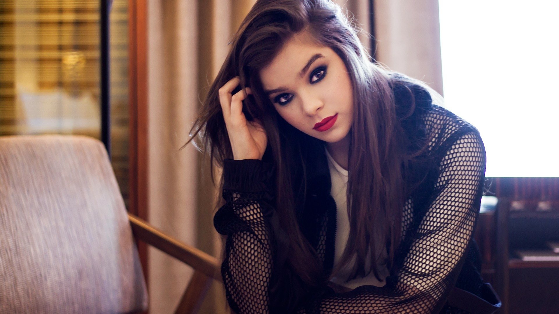 Brunette celebrity Hailee Steinfeld, singer and actress, poses indoors wearing bold lipstick and a stylish black mesh jacket.