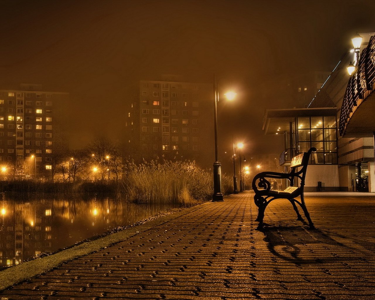 A solitary bench sits by the water's edge, illuminated by streetlights in a foggy urban setting, with buildings reflecting in the calm water.