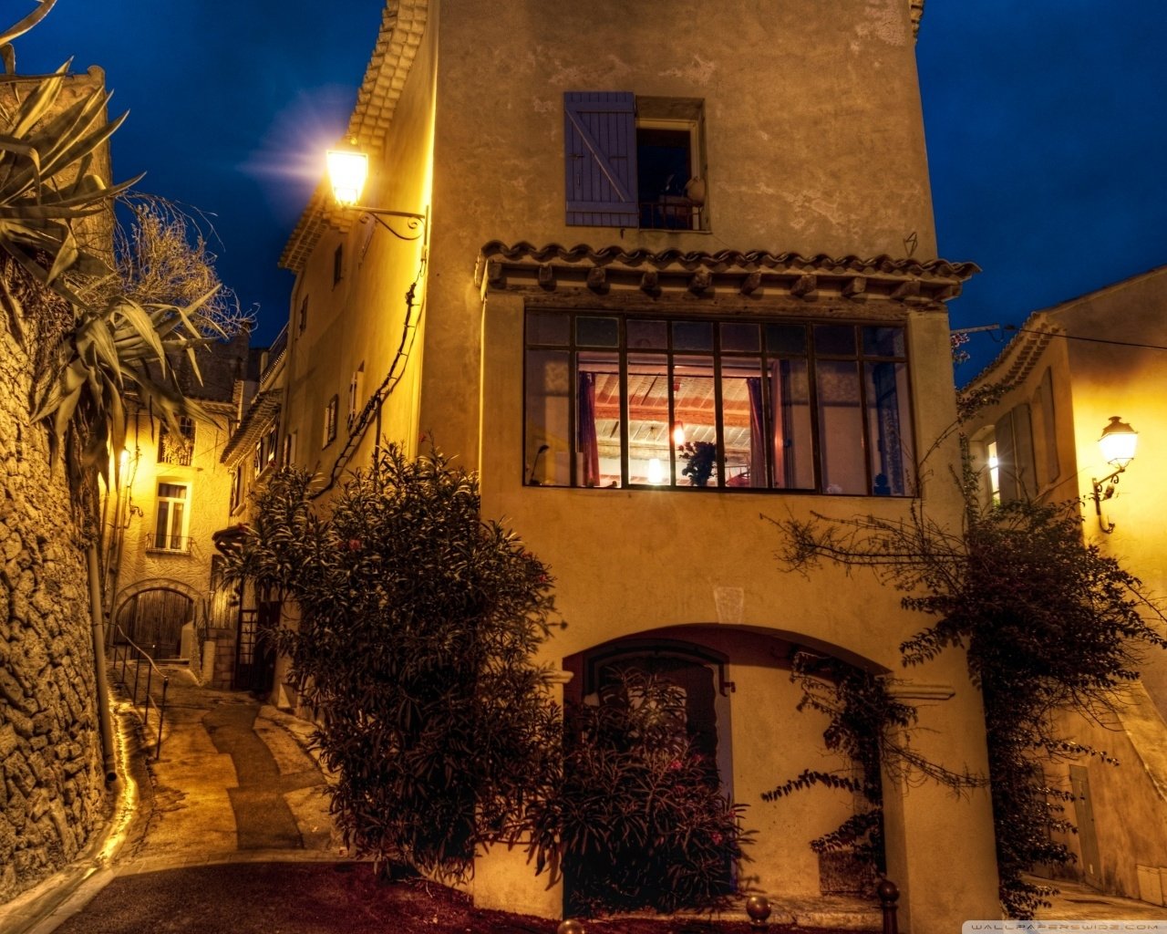 Warmly lit house facade at dusk on a narrow lane; man-made stucco walls, shuttered windows, street lanterns and climbing plants casting golden light.