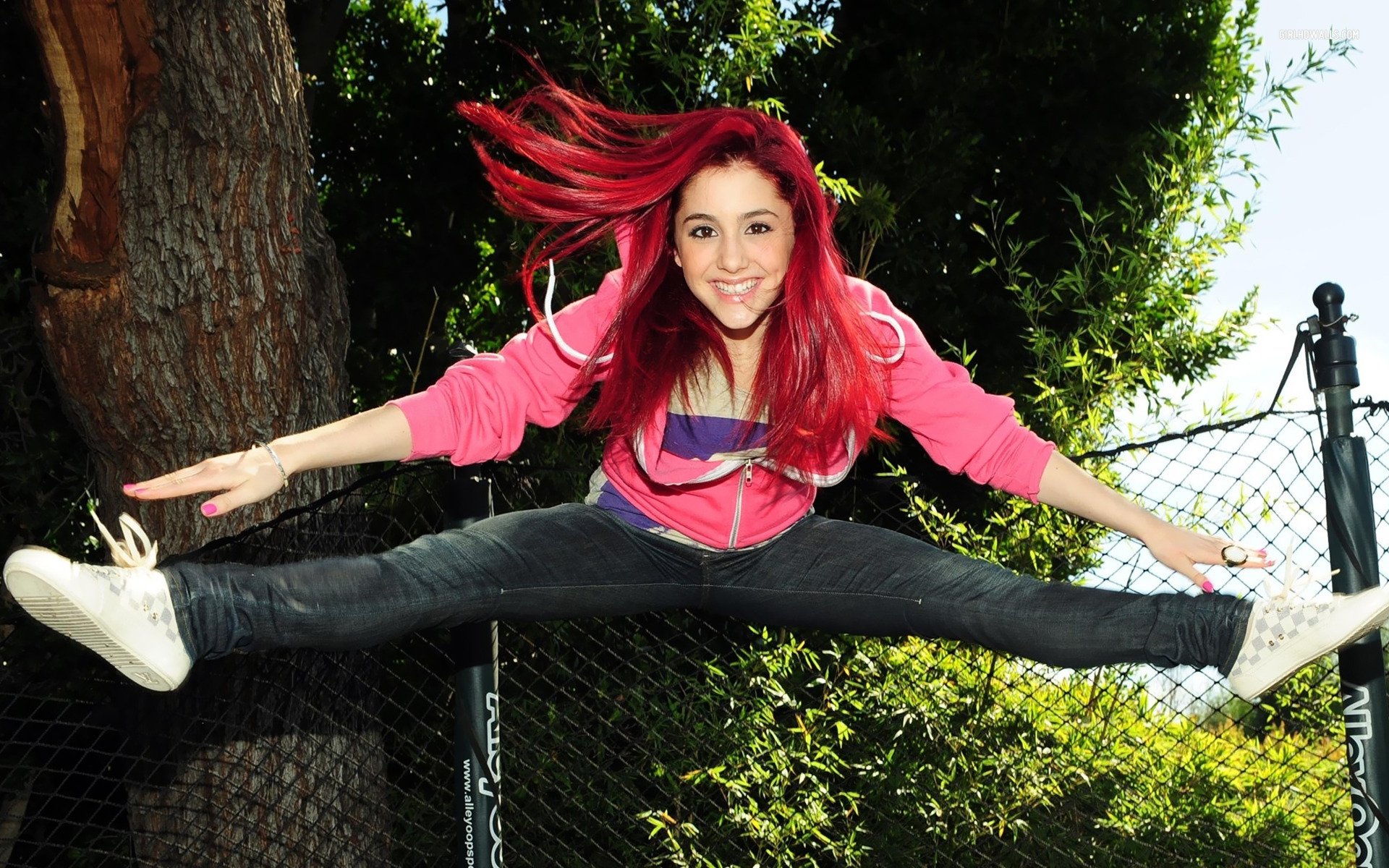 Ariana Grande jumps playfully on a trampoline, her vibrant red hair flowing as she stretches in mid-air, surrounded by greenery and a fence in the background.