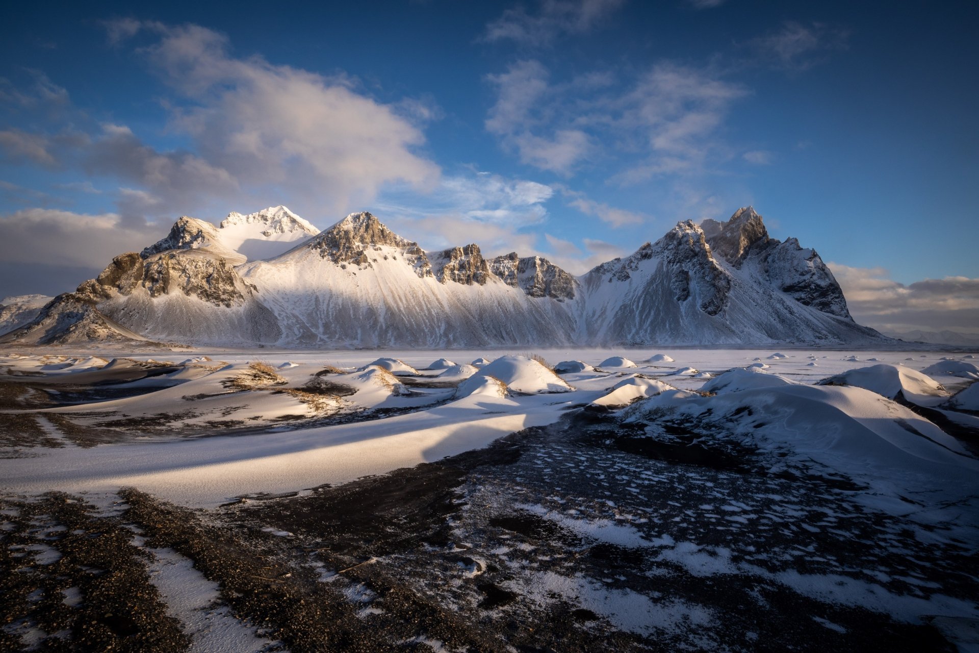 Vestrahorn Mountain in Iceland, snow-dusted jagged peaks rising above black sand dunes and shallow reflective pools under a dramatic blue sky — a stark nature landscape.