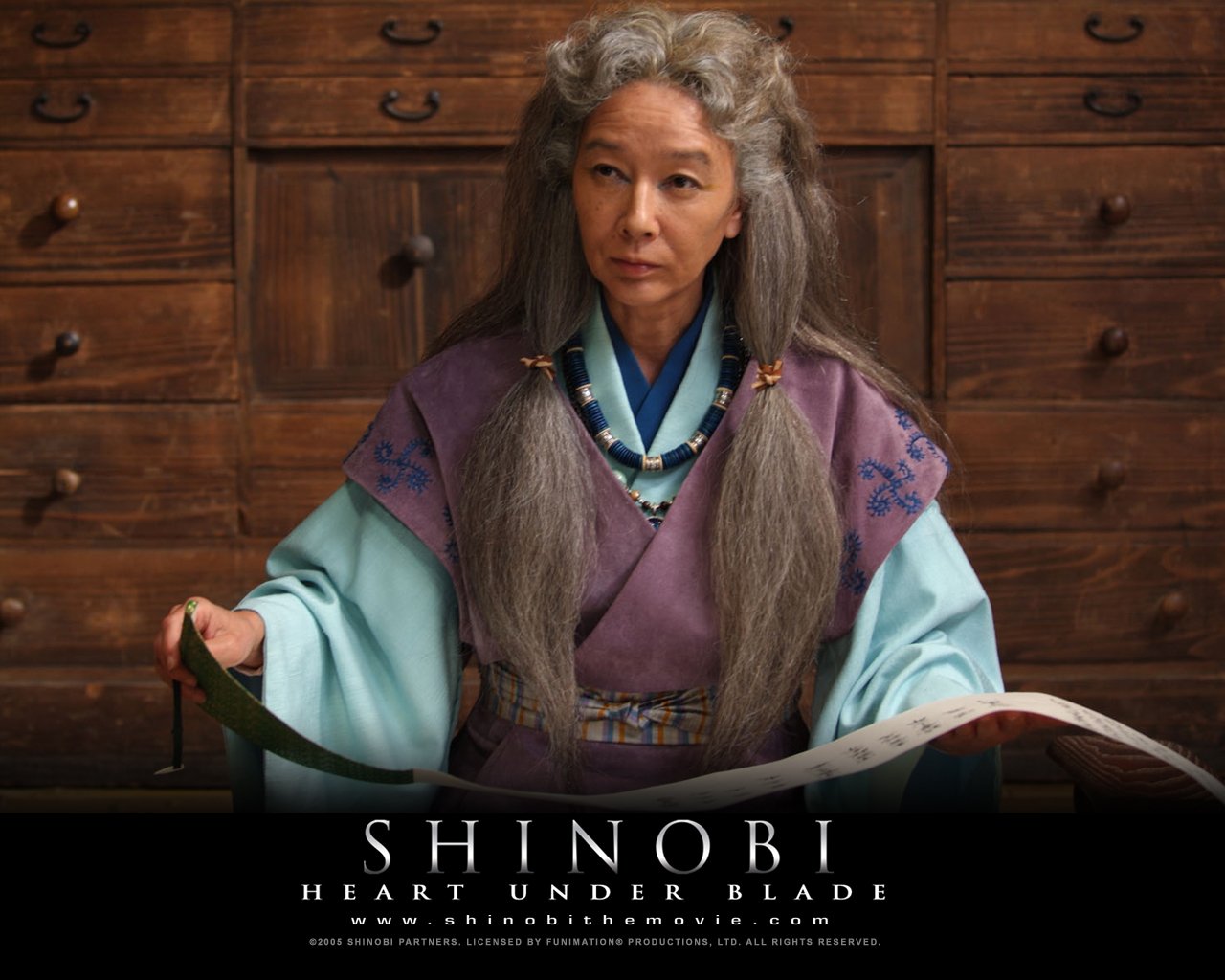 Elderly woman with long gray hair and traditional attire sits at a wooden desk in a scene from the movie Shinobi: Heart Under Blade.