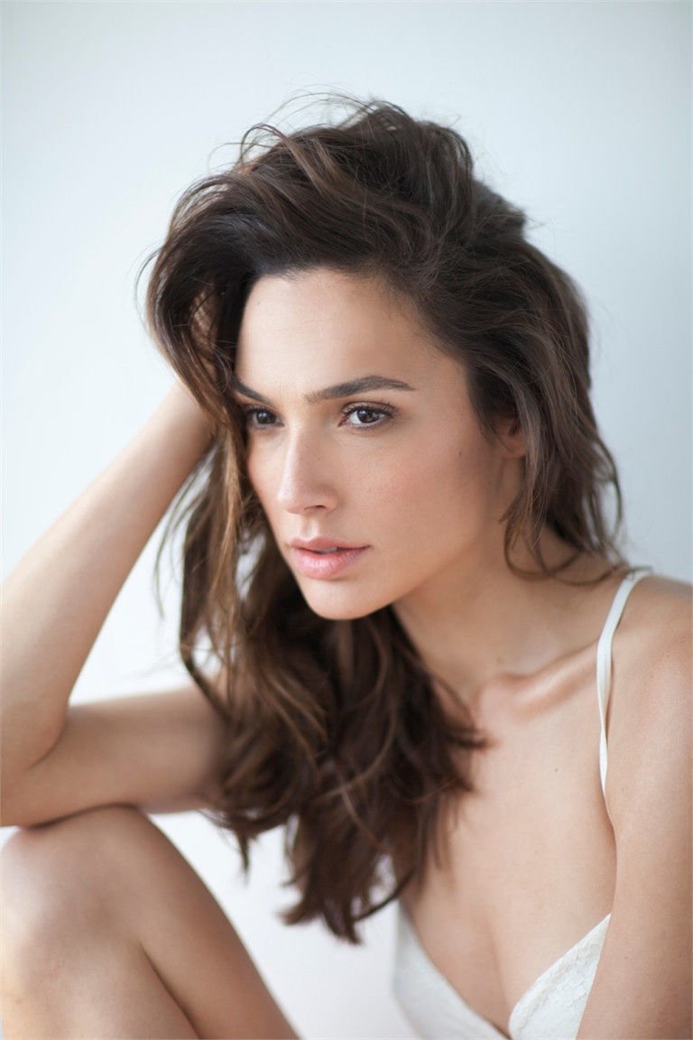 Close-up portrait of celebrity Gal Gadot with tousled hair, wearing a white top, gazing thoughtfully to the side against a light background.