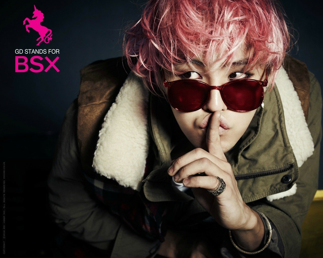 A stylish individual with pink hair and round sunglasses poses playfully with a finger to their lips. The image promotes music and features elements related to Bigbang.