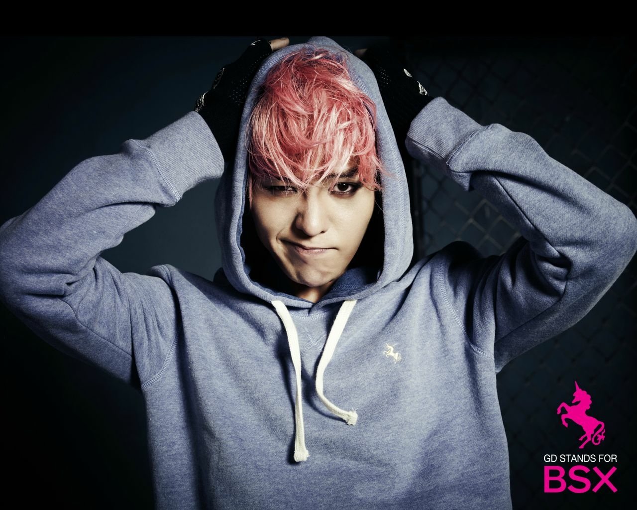 A young man with pink hair, wearing a gray hoodie, poses confidently against a dark background, embodying the vibrant energy of Bigbang's music scene.