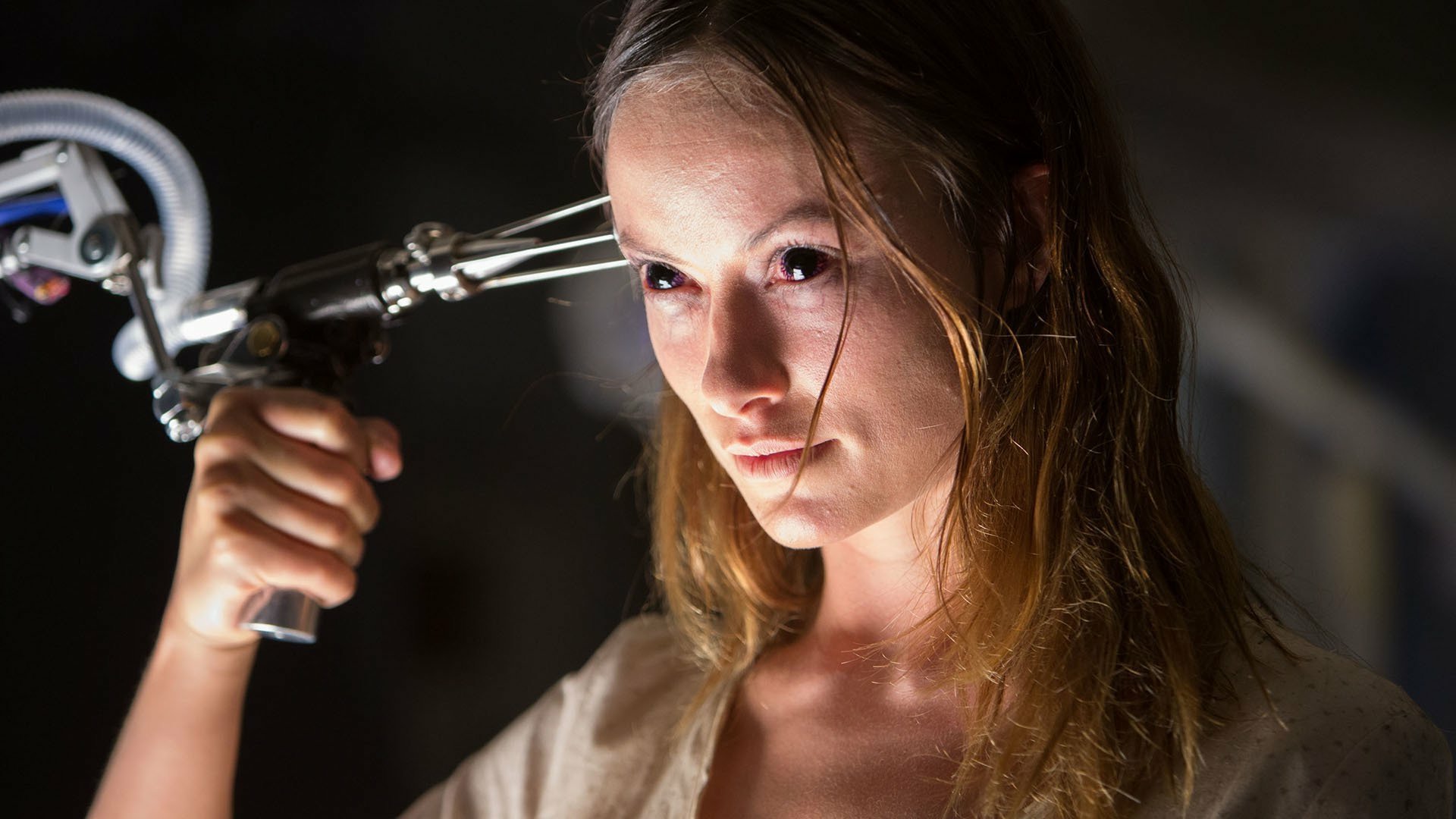 Actress from the movie The Lazarus Effect holds a surgical instrument beside her head, lit dramatically with a tense, focused expression.