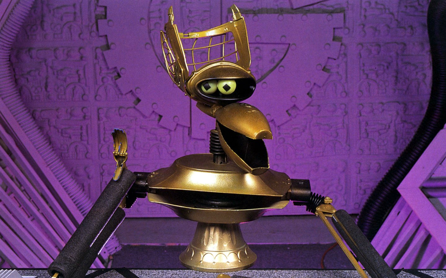 A robot character with a golden crown from the TV show Mystery Science Theater 3000, set against a purple backdrop with mechanical and geometric designs.