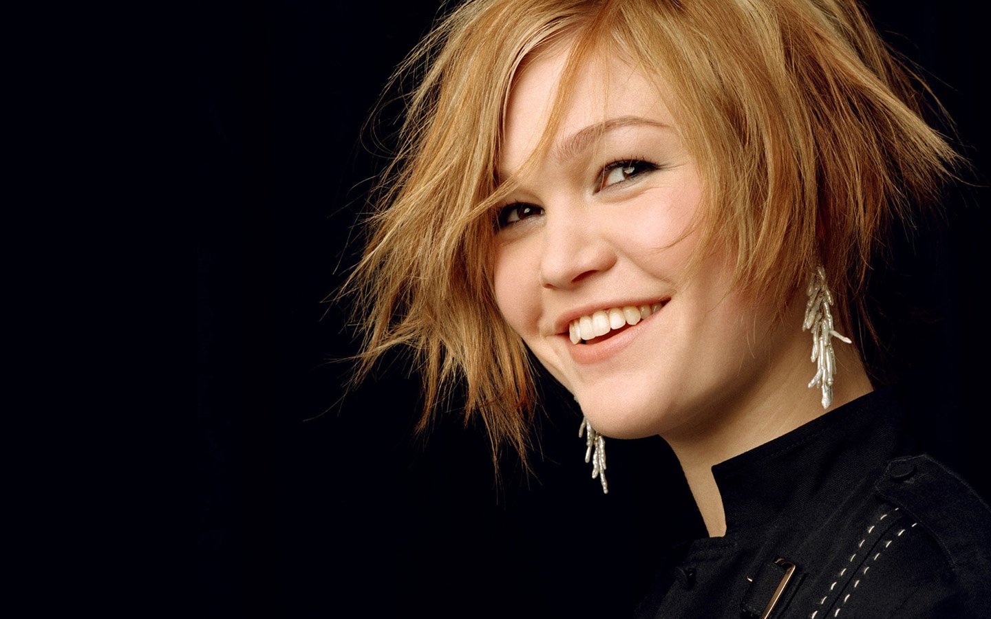 A smiling Julia Stiles showcases a playful short hairstyle, wearing statement earrings against a black background, radiating confidence and charm.
