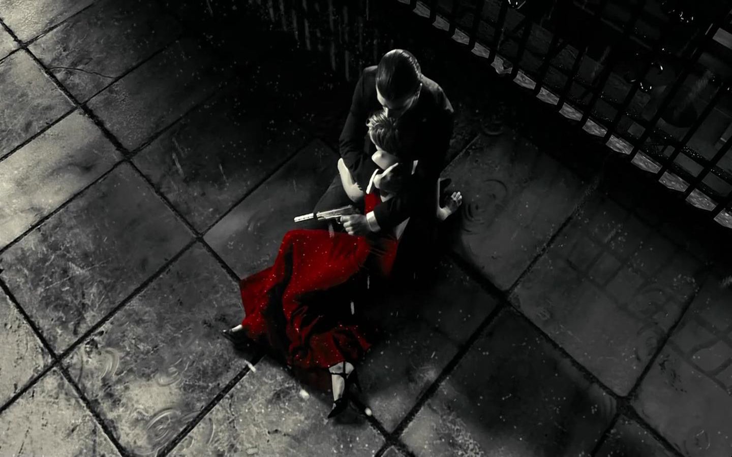 A dark, rain-soaked street from the movie Sin City shows a man holding an injured woman in a striking red dress.
