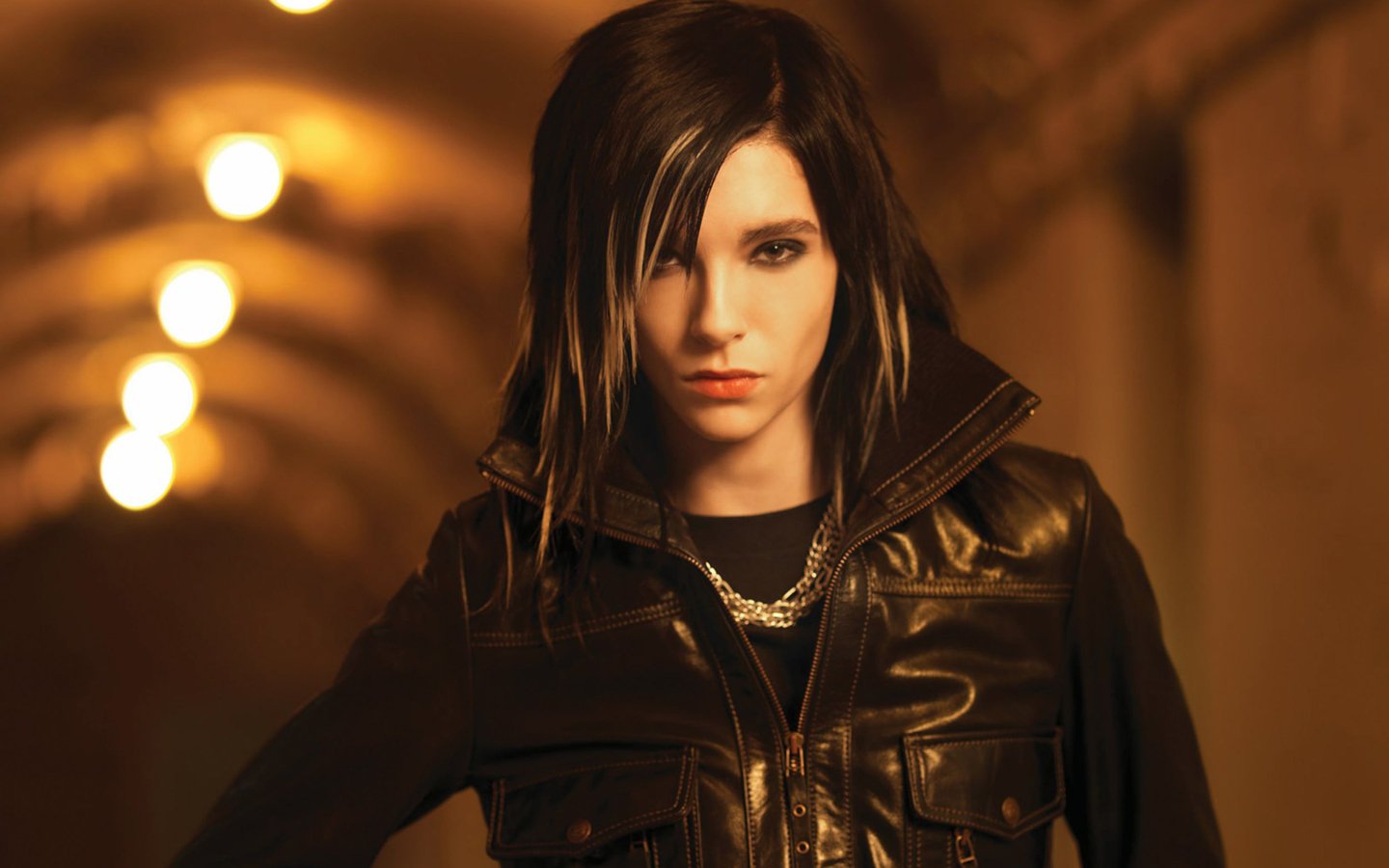 A musician from Tokio Hotel stands confidently in a dimly lit tunnel, wearing a black leather jacket with bold, expressive features and stylized hair.