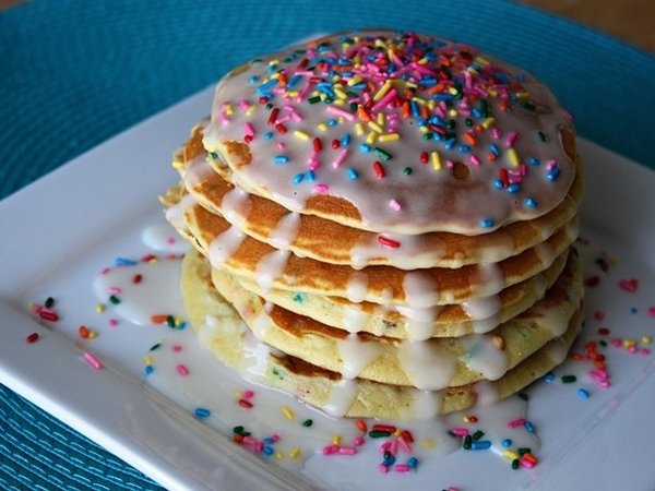 Stack of breakfast pancakes with white glaze and rainbow sprinkles, dessert-style food served on a white plate.