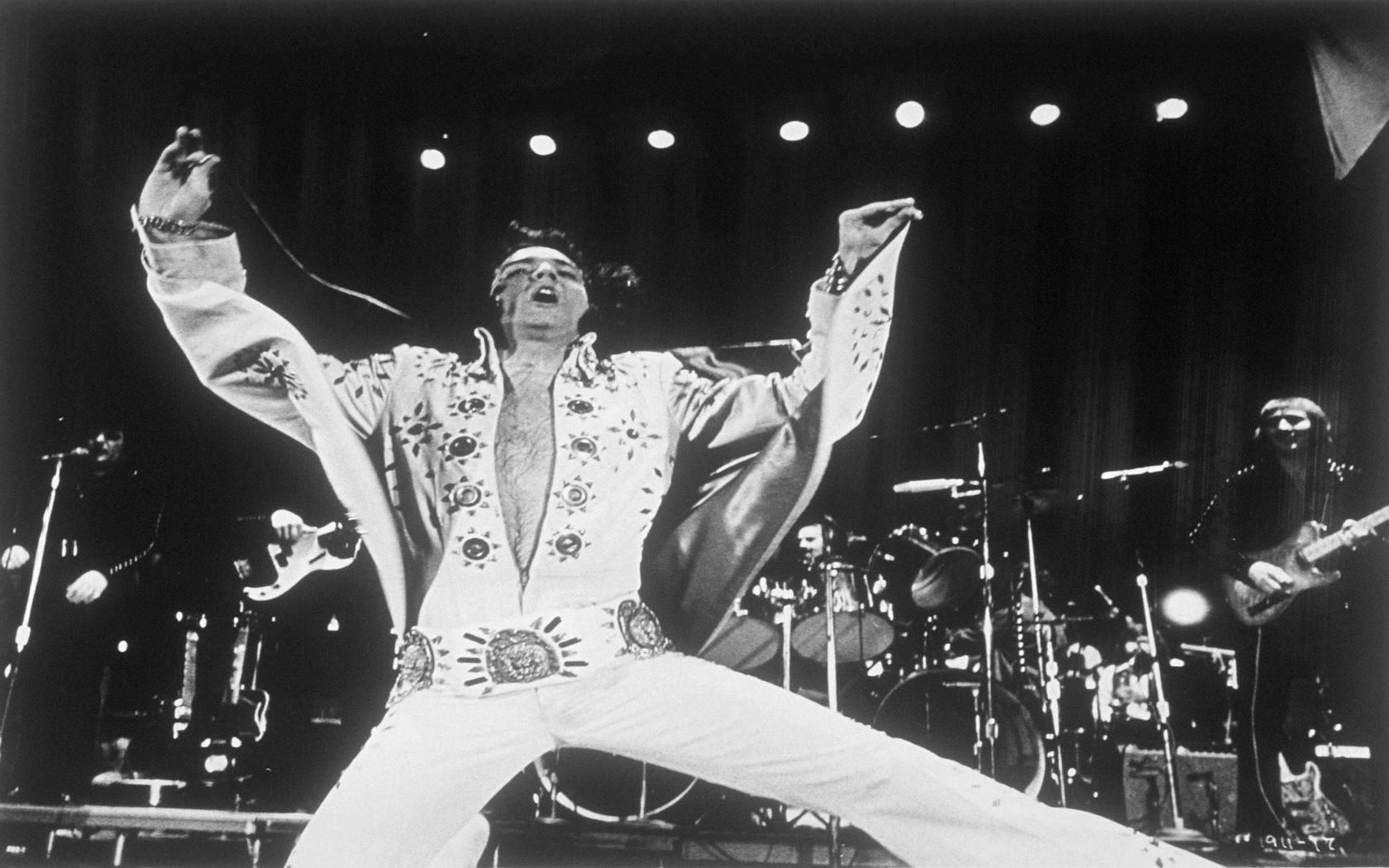 Elvis Presley performs energetically on stage in a white embellished jumpsuit, backed by his band under stage lights.