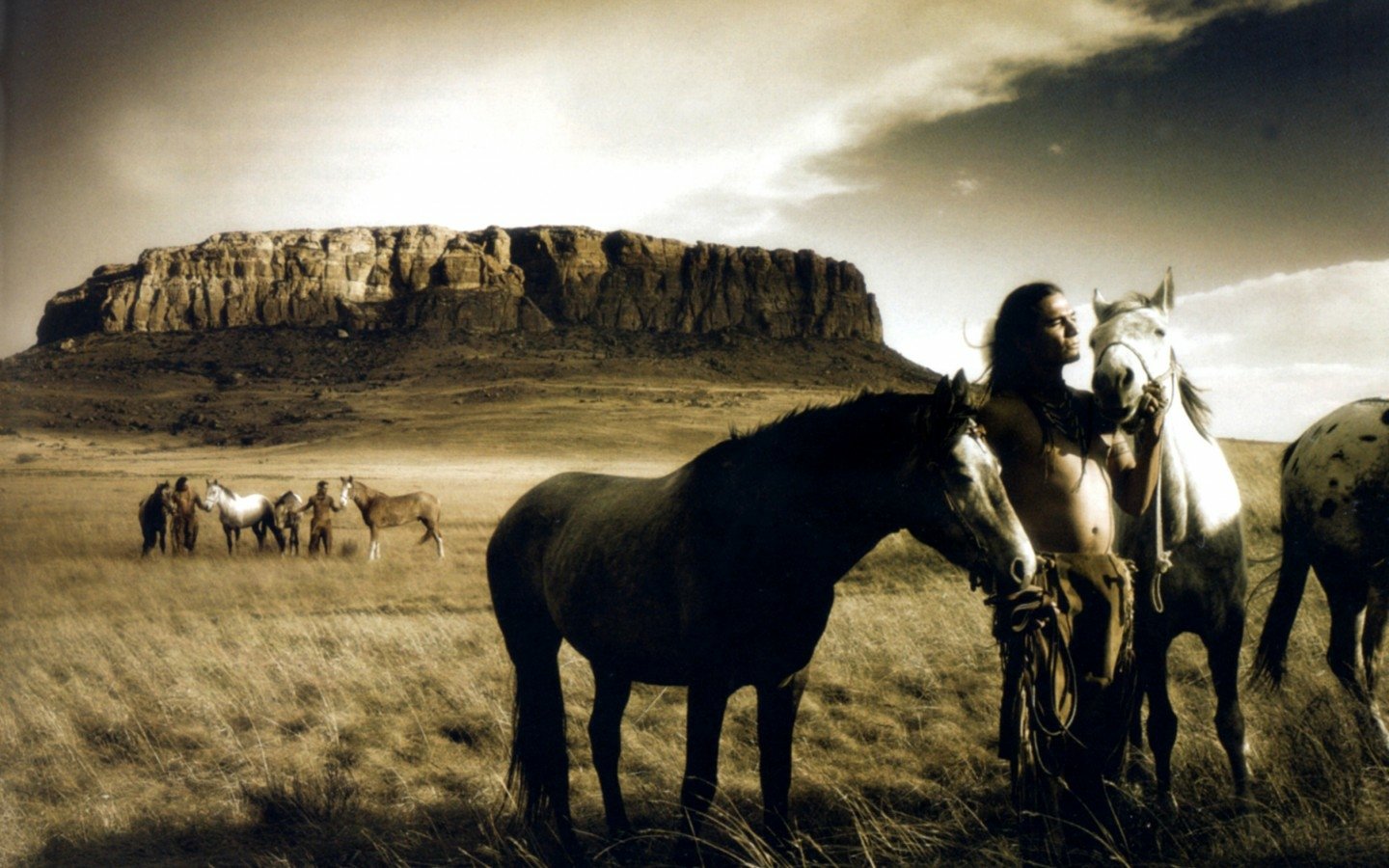 A Native American individual stands among horses in a sweeping landscape, with a dramatic mesa backdrop under a moody sky, capturing a connection to nature and heritage.