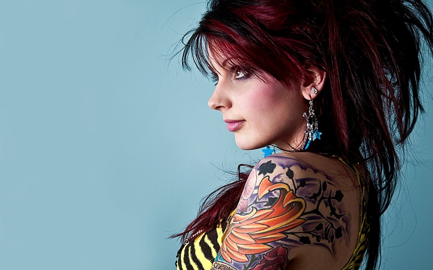 A woman with vibrant red and black hair poses against a light blue background, showcasing an intricate tattoo on her arm and stylish earrings.