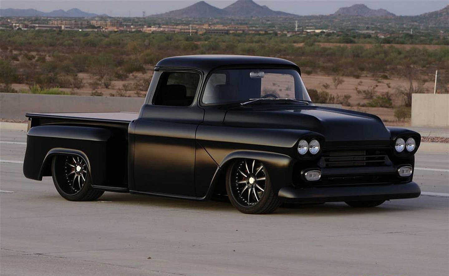 A sleek black truck parked on an open road, showcasing its classic design and modern wheels against a backdrop of desert mountains.