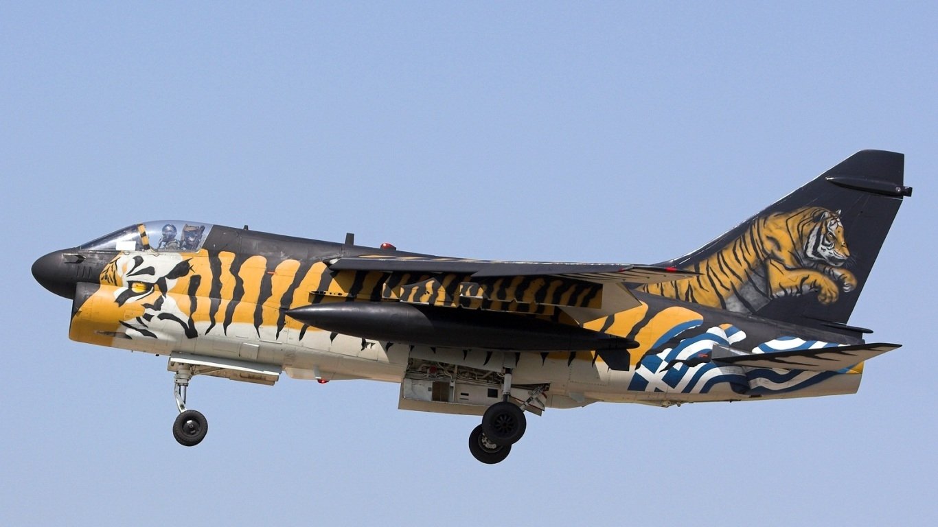 Military LTV A-7 Corsair II jet painted with bold tiger stripes and nose art, landing gear down against a clear blue sky.