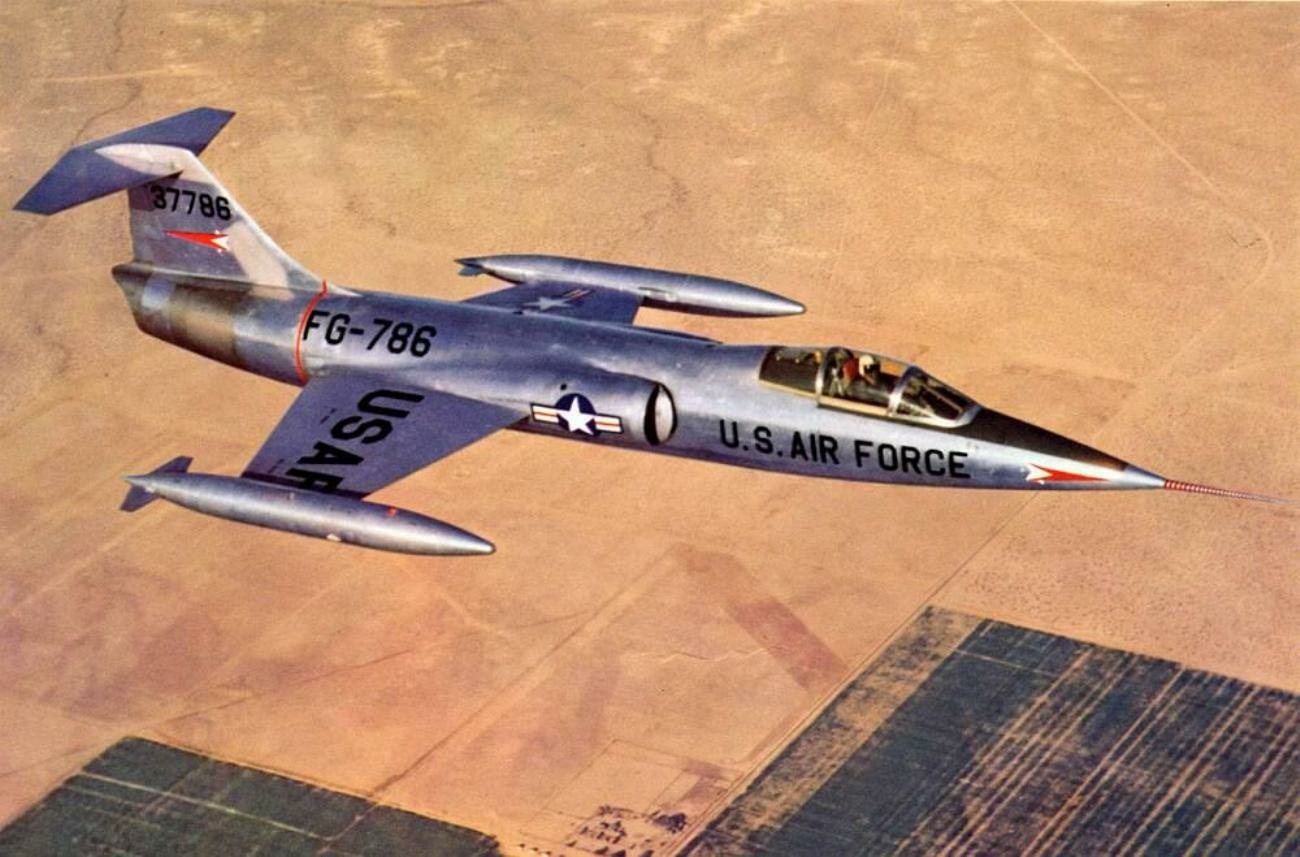 A U.S. Air Force Lockheed F-104 Starfighter jet flying over a patchwork of farmland and desert terrain.