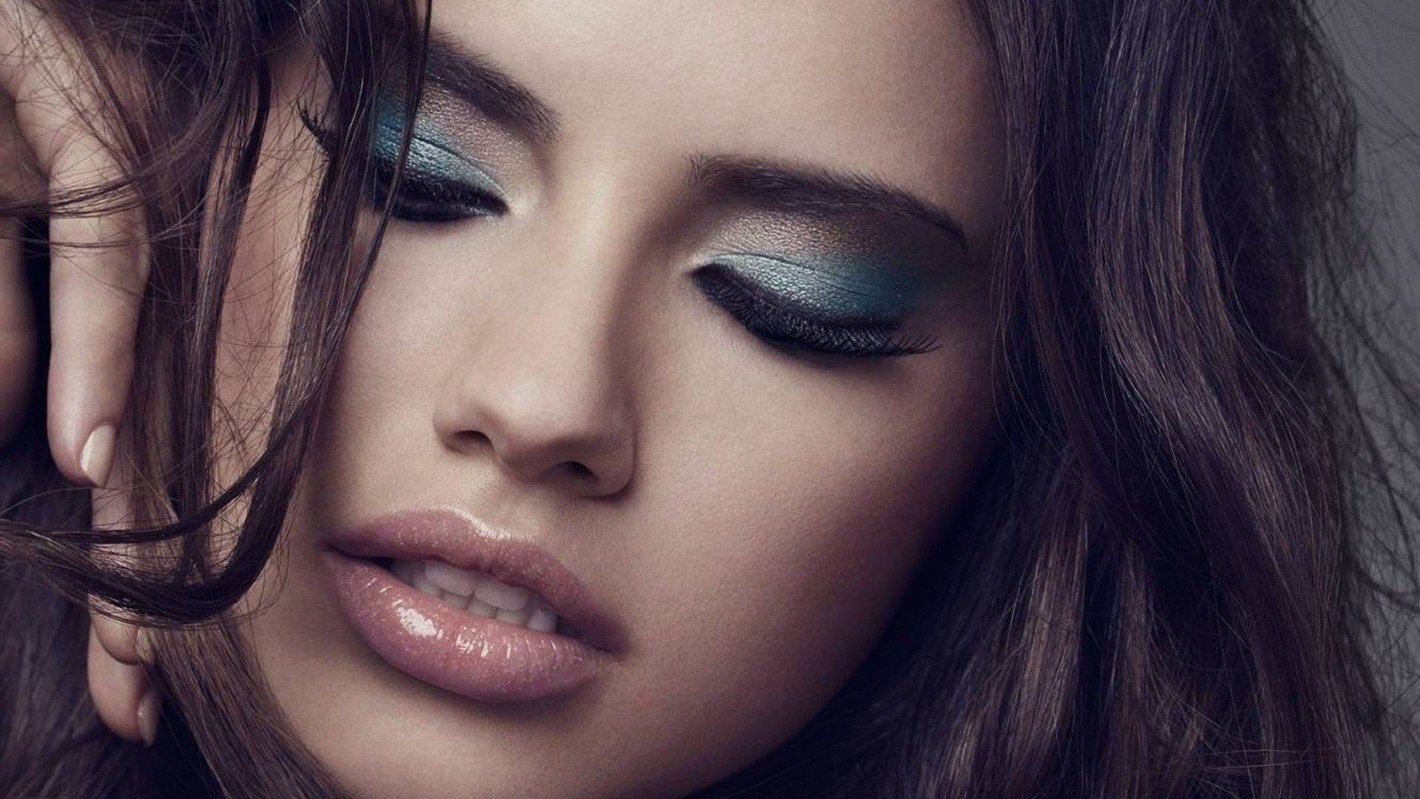 I can’t identify the person in the image. Alt: Close-up of a woman with glossy lips, teal smoky eyeshadow, long dark hair, eyes closed, hand near her face.