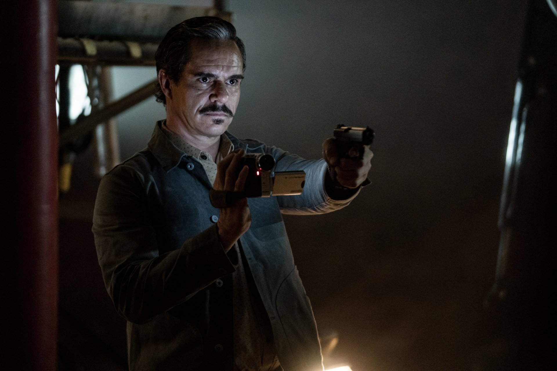 Lalo Salamanca from the TV show Better Call Saul holds a flashlight and aims a handgun in a dimly lit room, wearing a tense, focused expression.