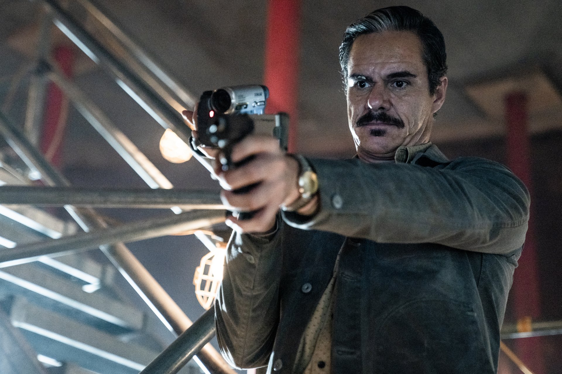 Lalo Salamanca from Better Call Saul aims a pistol at the viewer, grim expression and mustache framed by a dim, industrial stairwell backdrop.