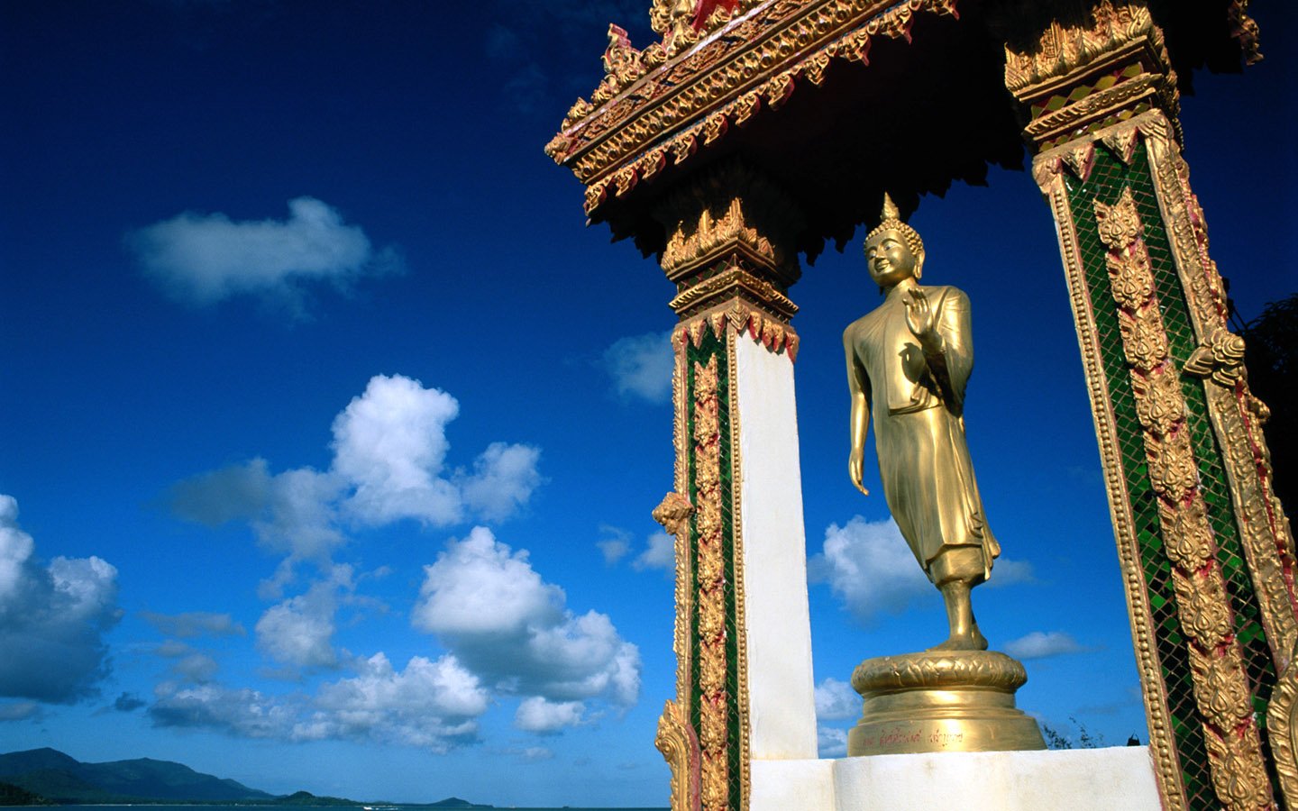 A golden Buddha statue stands gracefully under an ornate canopy, framed by a clear blue sky with fluffy clouds, symbolizing tranquility and spiritual peace in Buddhism.