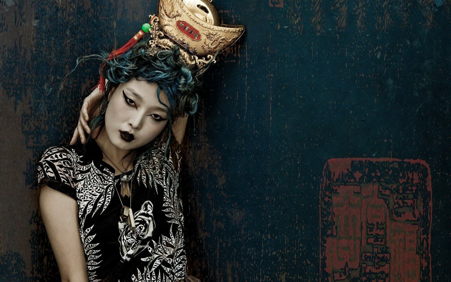 A woman styled as a geisha with elaborate makeup and a decorative mask resting on her head, posing against a dark textured background.