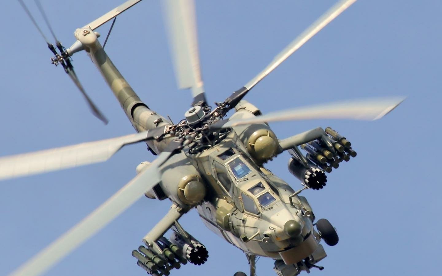 A military Mil Mi-28 attack helicopter in flight against a clear blue sky, showcasing its armament and rotor blades in motion.