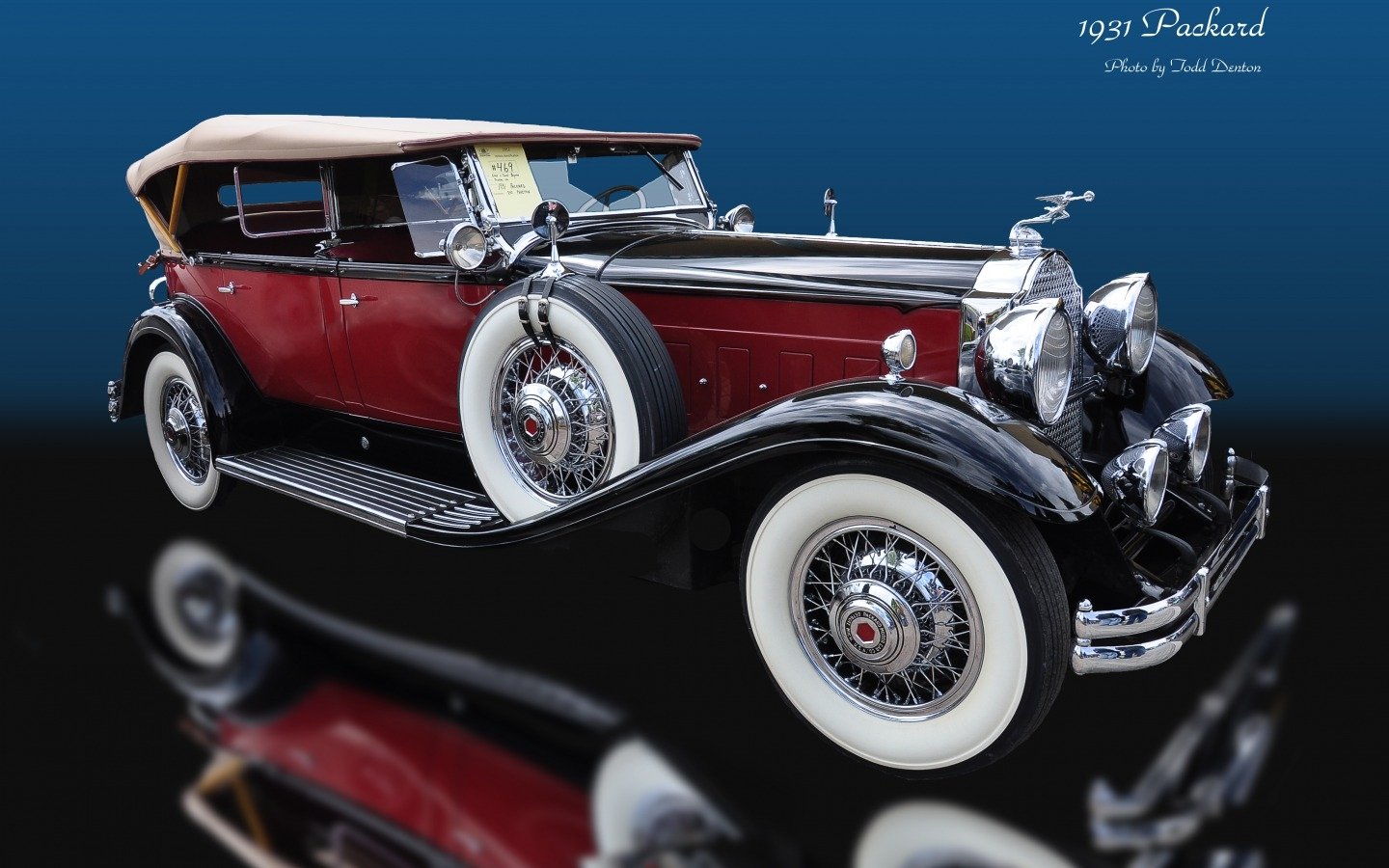1937 Packard vehicle: maroon and black vintage convertible with tan soft top, whitewall tires, chrome headlights and ornate grille, reflected on a glossy black surface.