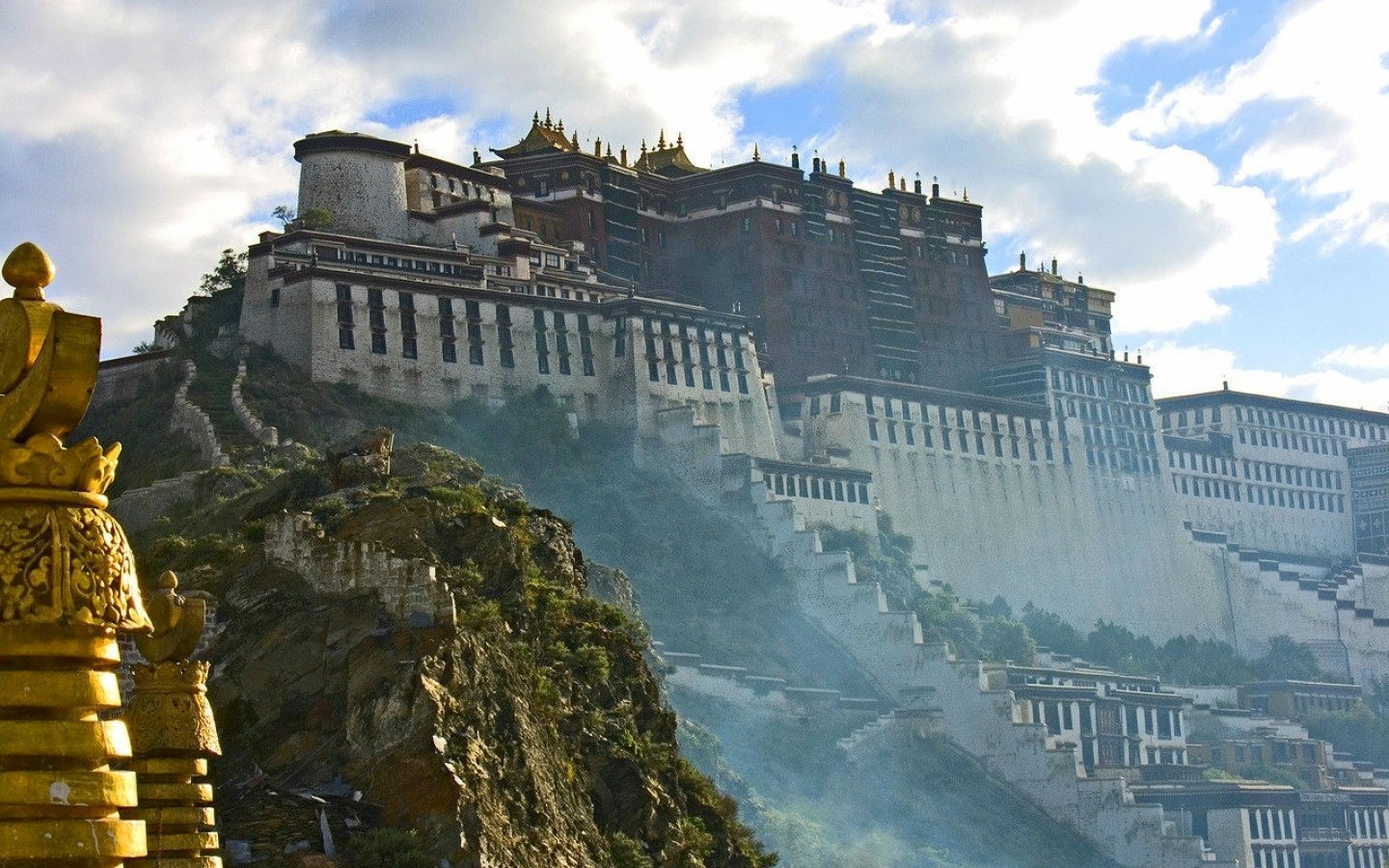 The Potala Palace, a grand man-made structure, rises majestically on a hillside under a partly cloudy sky.