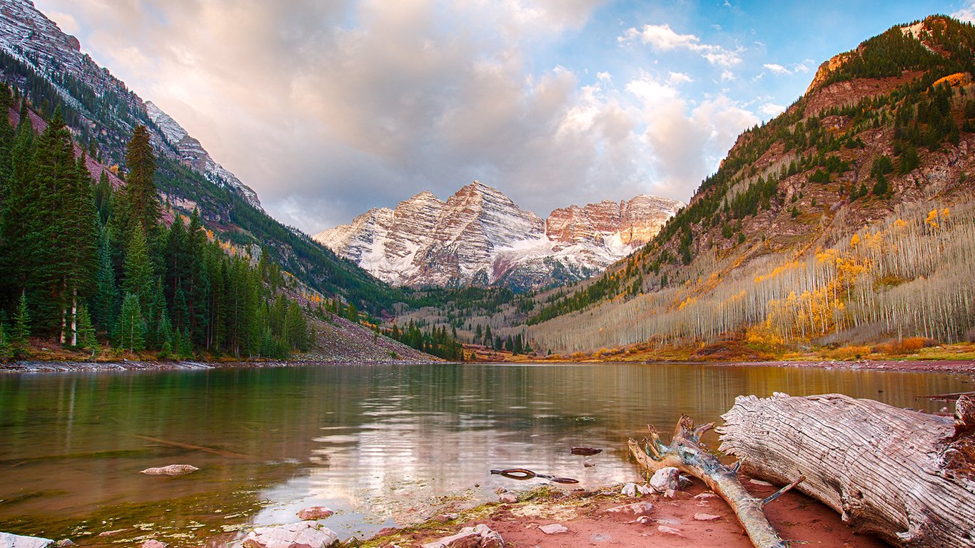A tranquil mountain lake surrounded by towering peaks and dense evergreen forests under a partly cloudy sky at sunset.