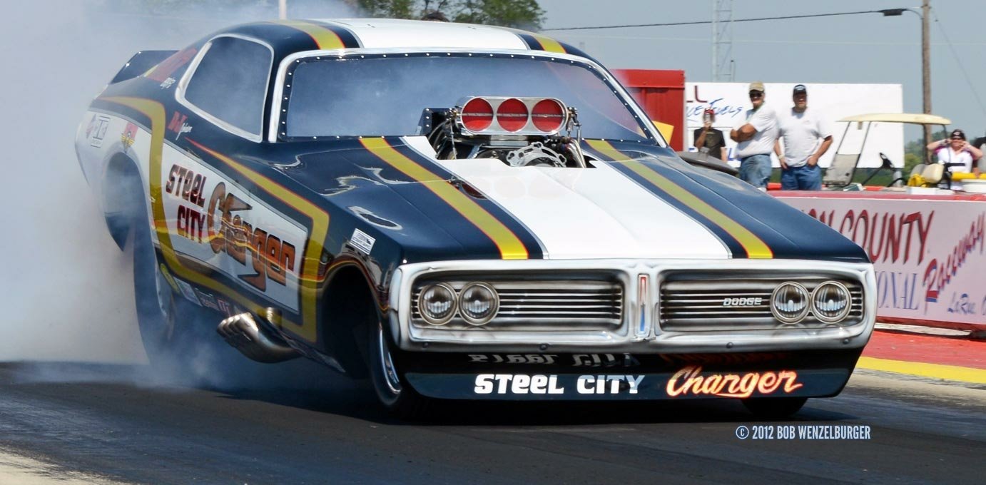 Blue-and-white Dodge Charger drag car with exposed supercharger, front wheels lifted as it launches into a smoke-filled burnout on the race strip.