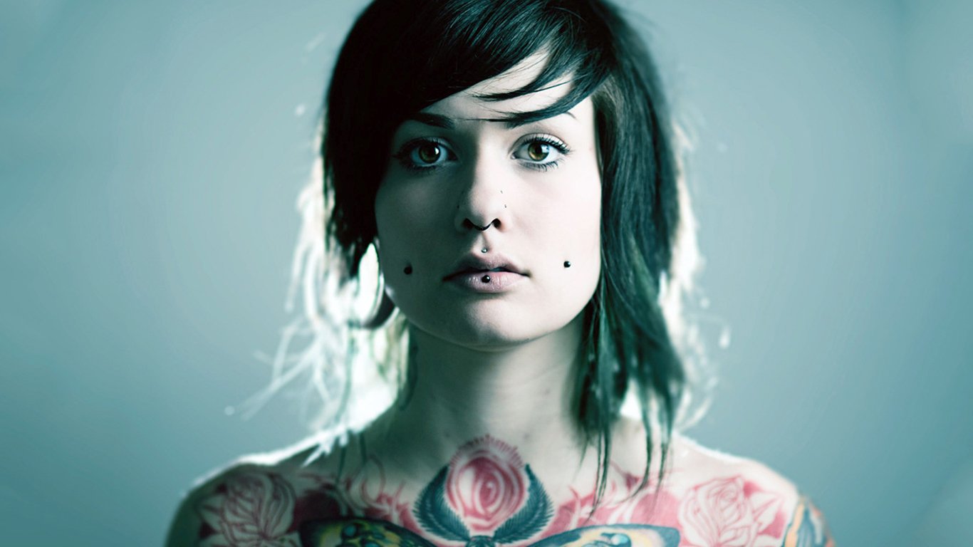 A portrait of a woman with striking tattoos, featuring colorful floral designs on her chest and a thoughtful expression, set against a soft blue background.