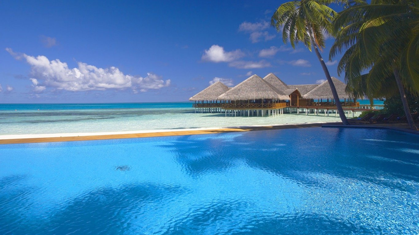 A tranquil scene of a tropical hut over the blue ocean in the Maldives, framed by palm trees and a shimmering pool, capturing the essence of a serene holiday getaway.