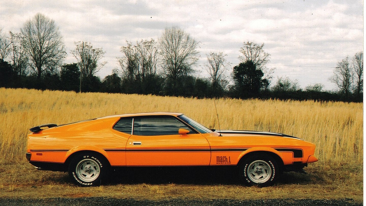 1972 Ford Mustang Mach 1 in bright orange parked on a rural dirt path with dry grass and leafless trees in the background.