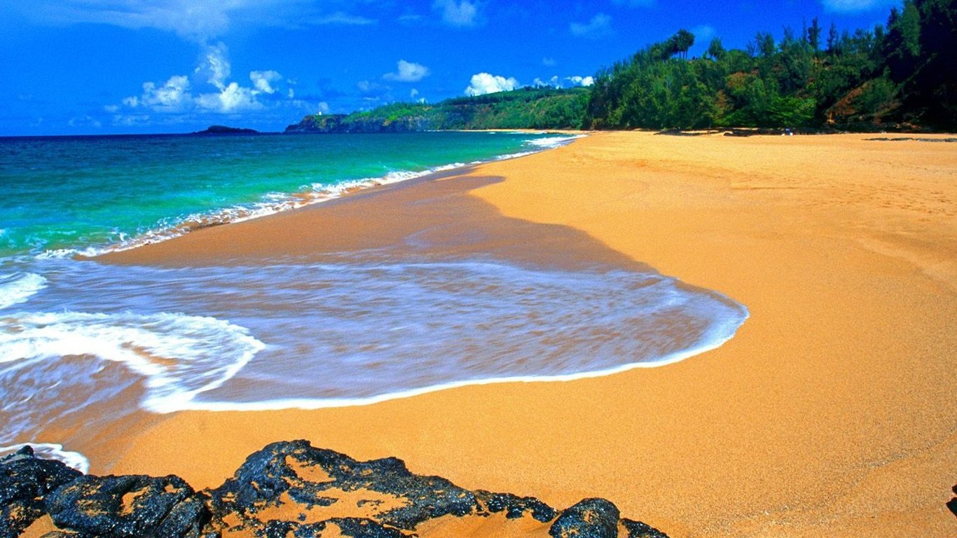 Golden sandy beach on Kauai, Hawaii with turquoise Pacific waves, rocky shoreline and lush green hills beneath a bright blue sky.