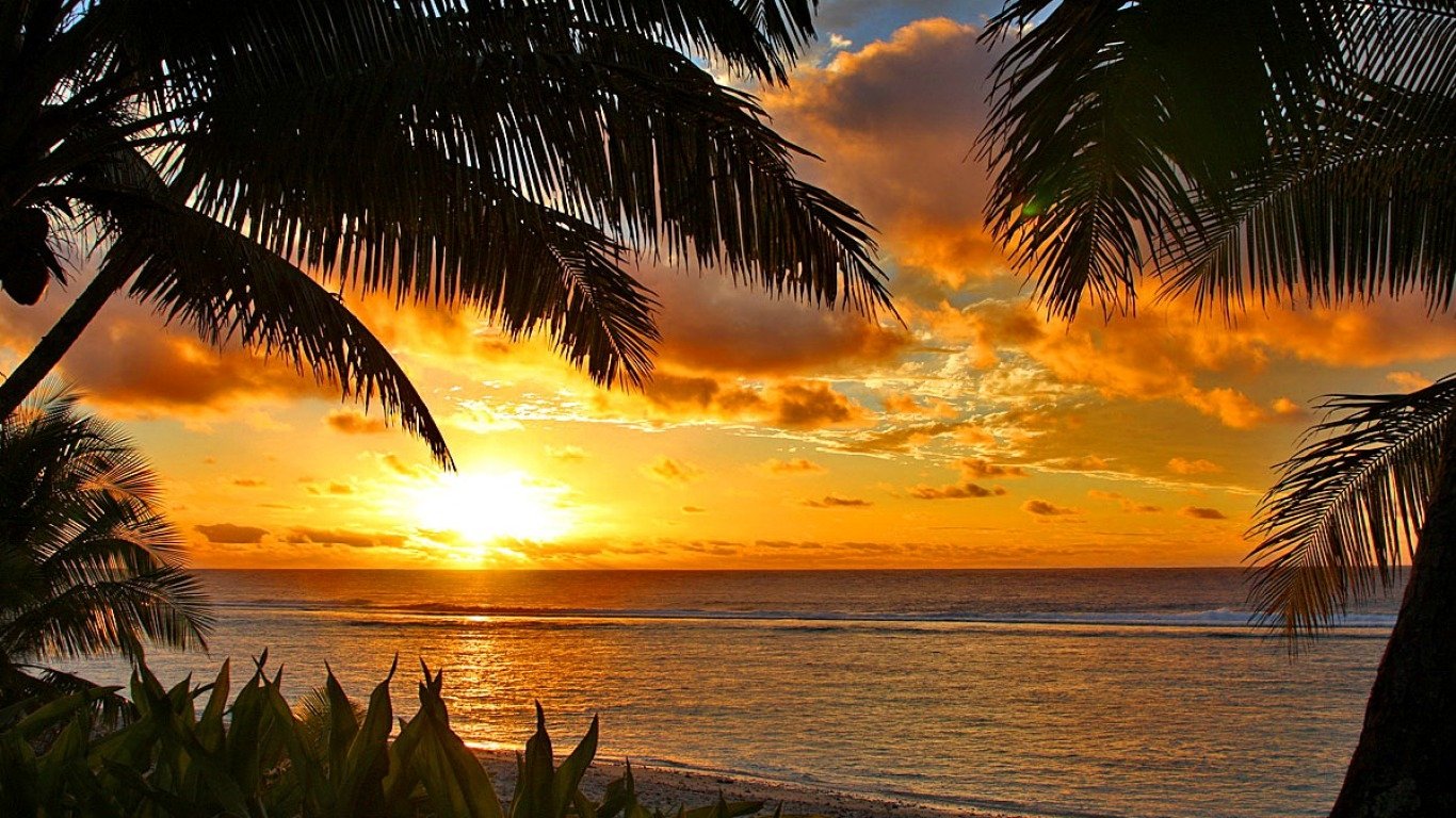 A vibrant sunset over the ocean, framed by tropical palm trees, creates a serene beach scene, showcasing the beauty of nature. Warm hues reflect off the water, enhancing the tranquil atmosphere.