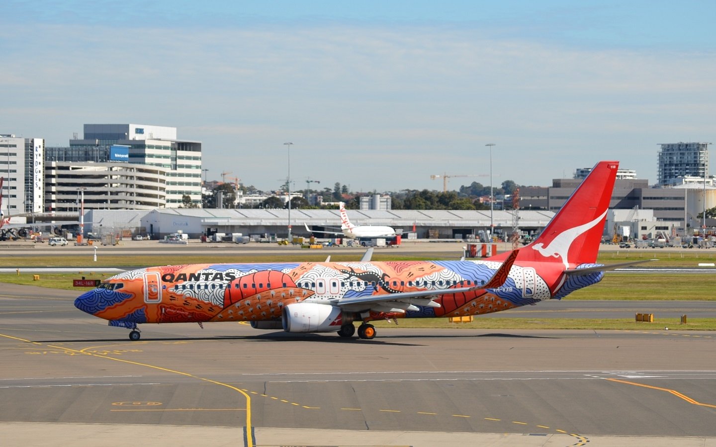 A Qantas Boeing 737 aircraft featuring the Yananyi Dreaming livery taxis on an airport runway, with city buildings visible in the background.