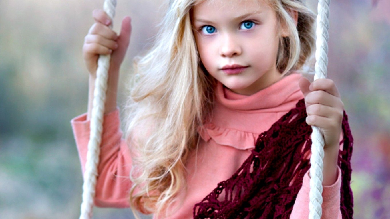 A young child with long, wavy blonde hair sits on a swing, gazing thoughtfully at the camera, dressed in a pink top and a burgundy shawl against a softly blurred background.
