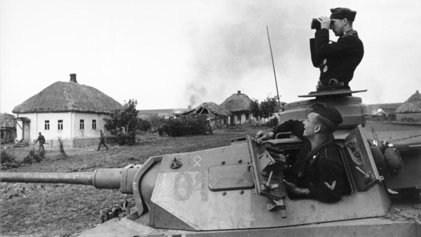 Black-and-white military photo of a Sturmgeschütz III with crew; one man in the cupola scanning with binoculars beside a rural village of thatched-roof houses.