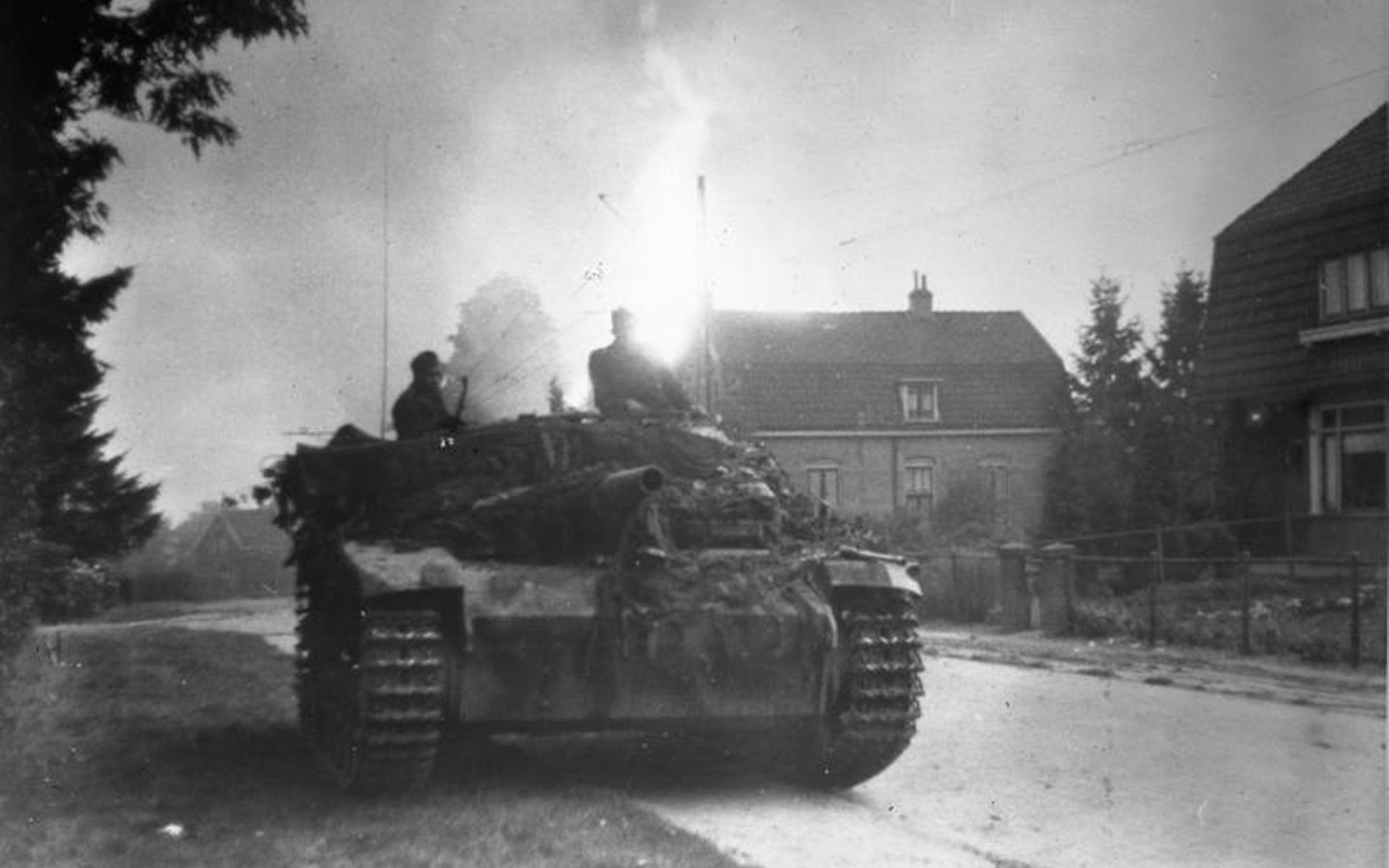 Sturmgeschütz III assault gun with crew advancing down a village street, camouflaged and raising smoke, houses visible in the background.