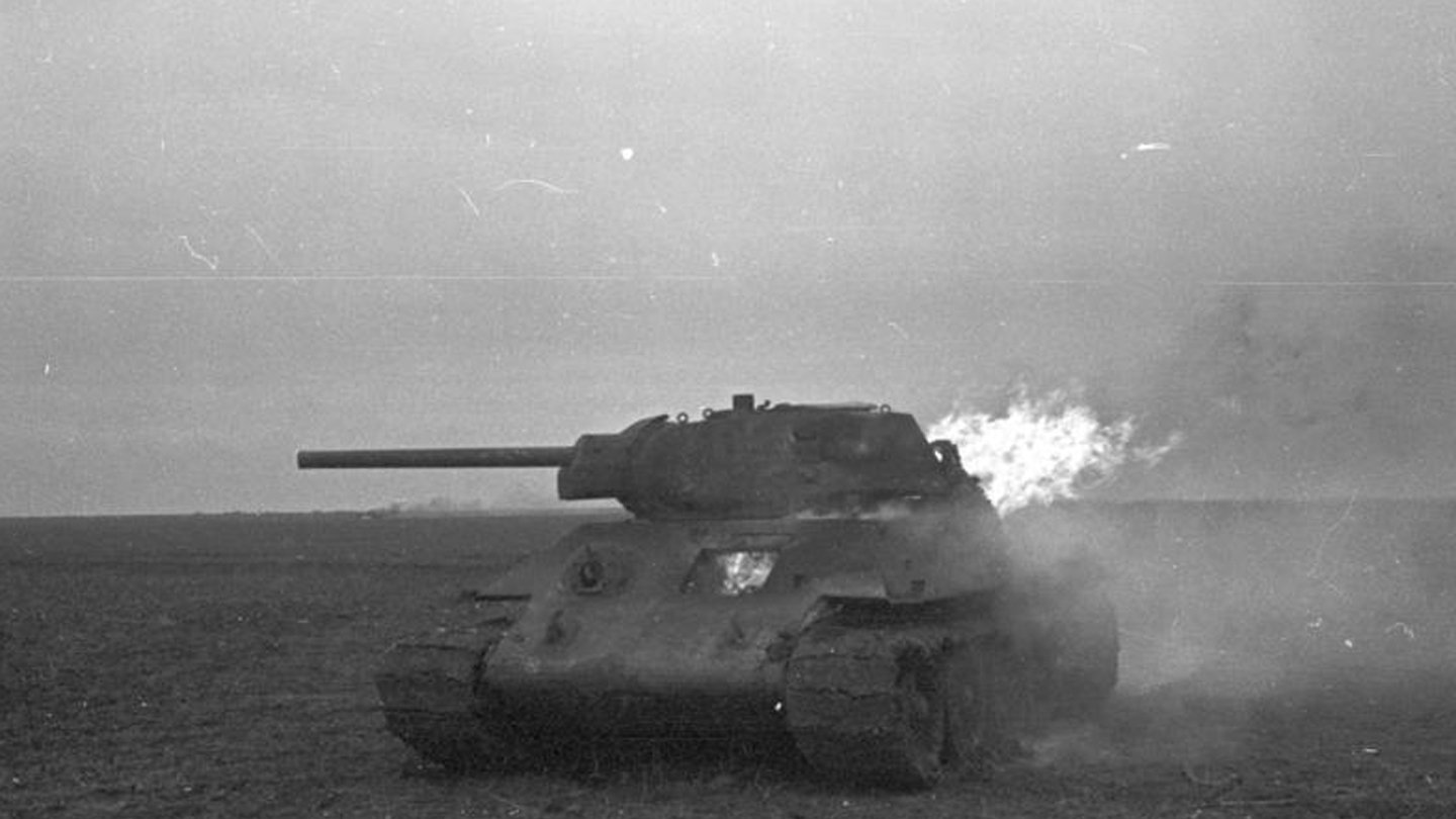 Black and white photo of a T-34 military tank firing its main gun in an open field, with visible smoke and flame from the barrel.